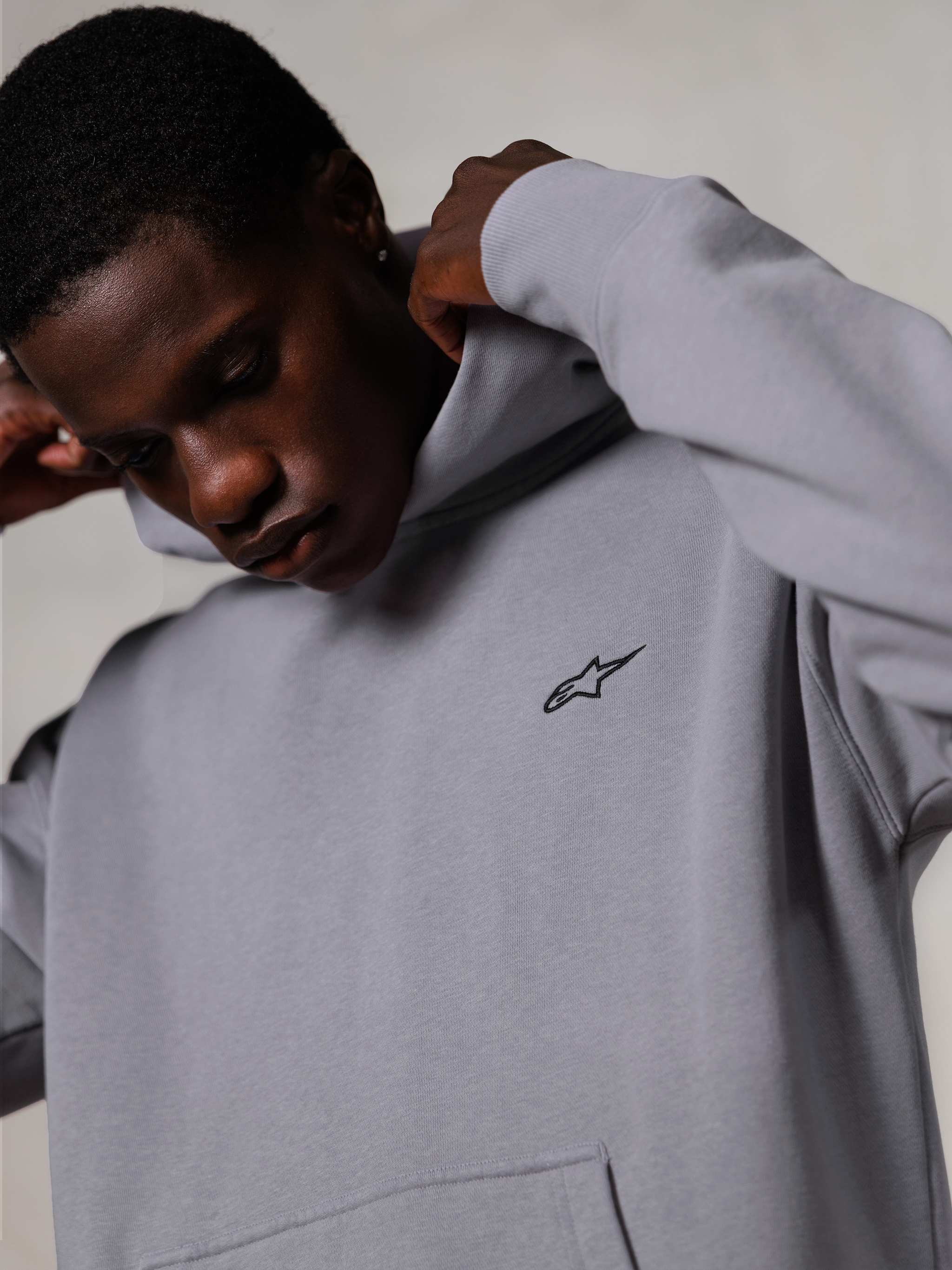 Alpinestars Essentials Ageless Hoodie, Silver, grande plano de um homem a ajustar o capuz da sweatshirt cinzenta, pequeno logótipo preto Alpinestars no peito, vestuário casual premium inspirado no desporto motorizado