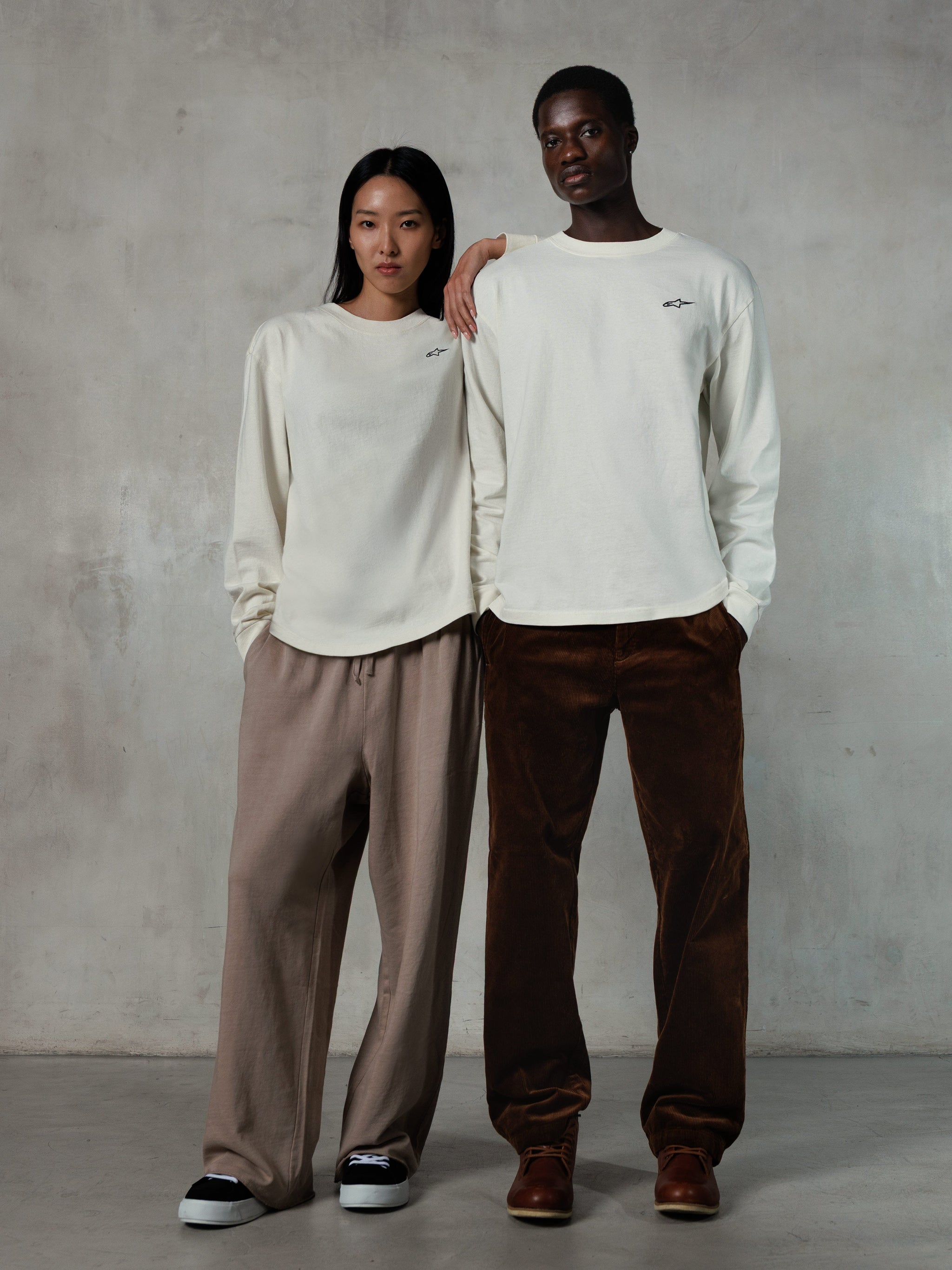 Alpinestars Essentials Ageless LS Tee, t-shirt de manga comprida, Off White, vestida por modelos masculino e feminino, pequeno logótipo preto da Alpinestars no peito, corte relaxado, combinada com calças de fato de treino beges e calças de bombazina castanhas, streetwear casual inspirado no desporto motorizado