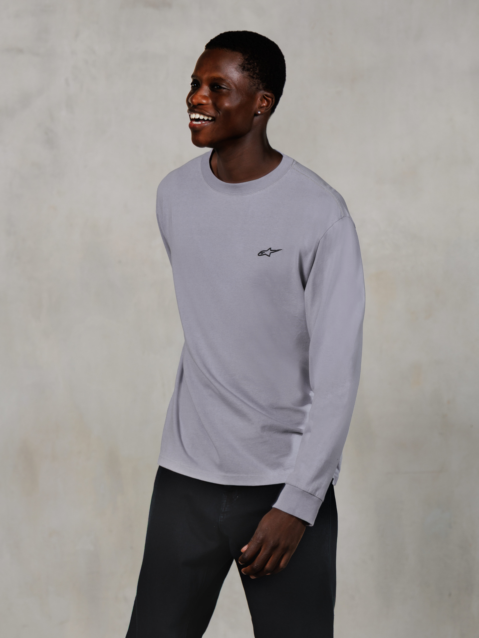 Alpinestars Essentials Ageless LS Tee, t-shirt de manga comprida prateada, pequeno logótipo preto no peito, vestida por um modelo a sorrir com calças escuras, ajuste contemporâneo descontraído, vestuário casual de estilo de vida motorsport