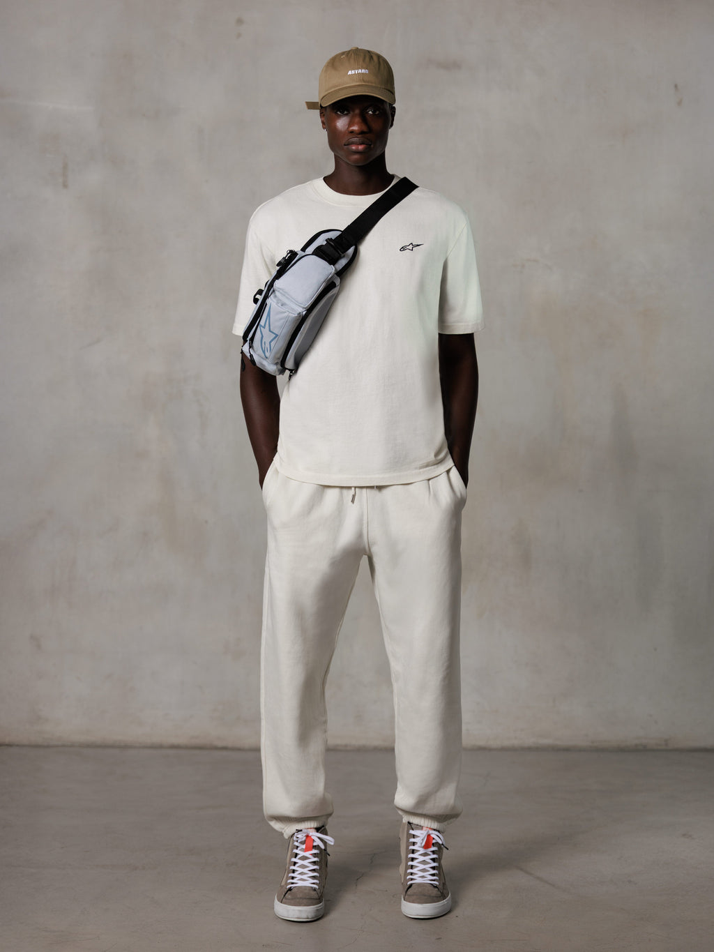 Alpinestars Essentials Ageless SS Tee, t-shirt de manga curta, Off White, com um pequeno logótipo preto no peito, usada com calças jogger a condizer, boné de basebol bege e mala a tiracolo cinzenta, estilo casual lifestyle streetwear