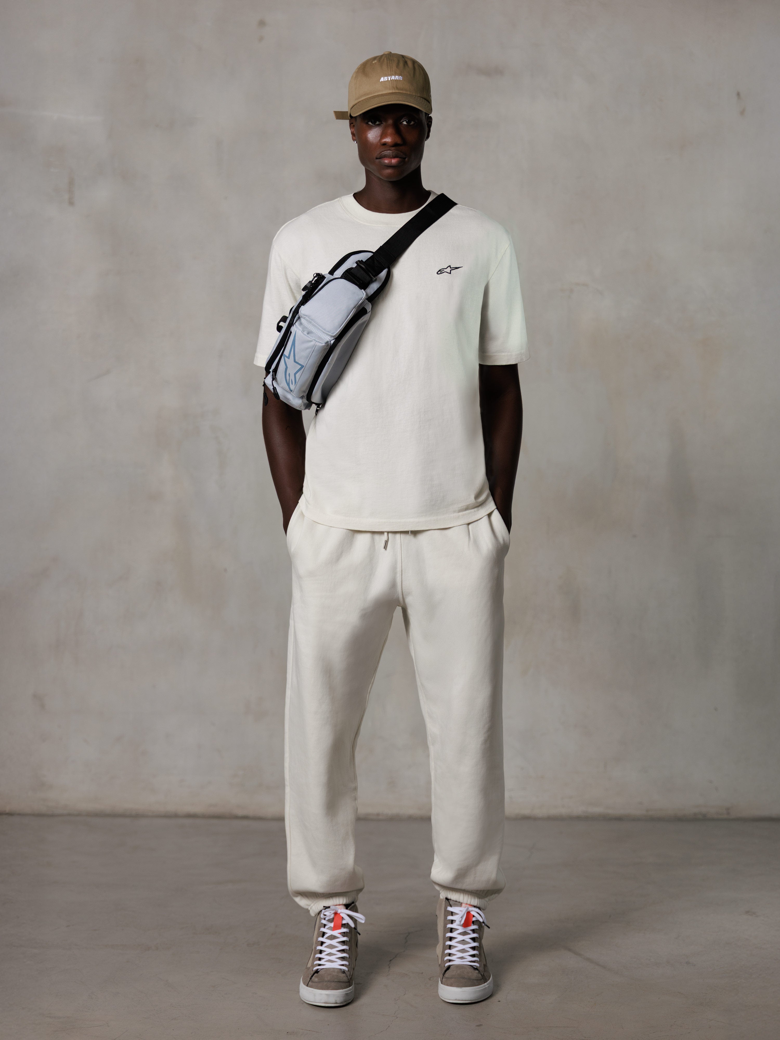 Alpinestars Essentials Ageless SS Tee, t-shirt de manga curta, Off White, com um pequeno logótipo preto no peito, usada com calças jogger a condizer, boné de basebol bege e mala a tiracolo cinzenta, estilo casual lifestyle streetwear