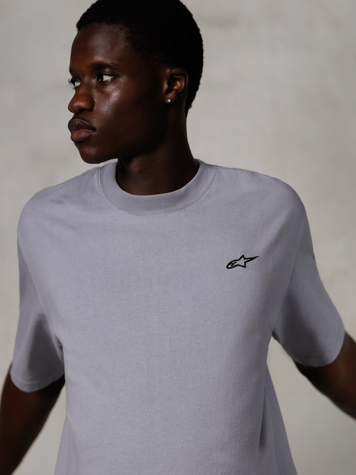 Modelo a usar Alpinestars Essentials Ageless SS Tee, t-shirt de manga curta, prateada, com um pequeno logótipo preto no peito, corte relaxado, design de streetwear simples e minimalista