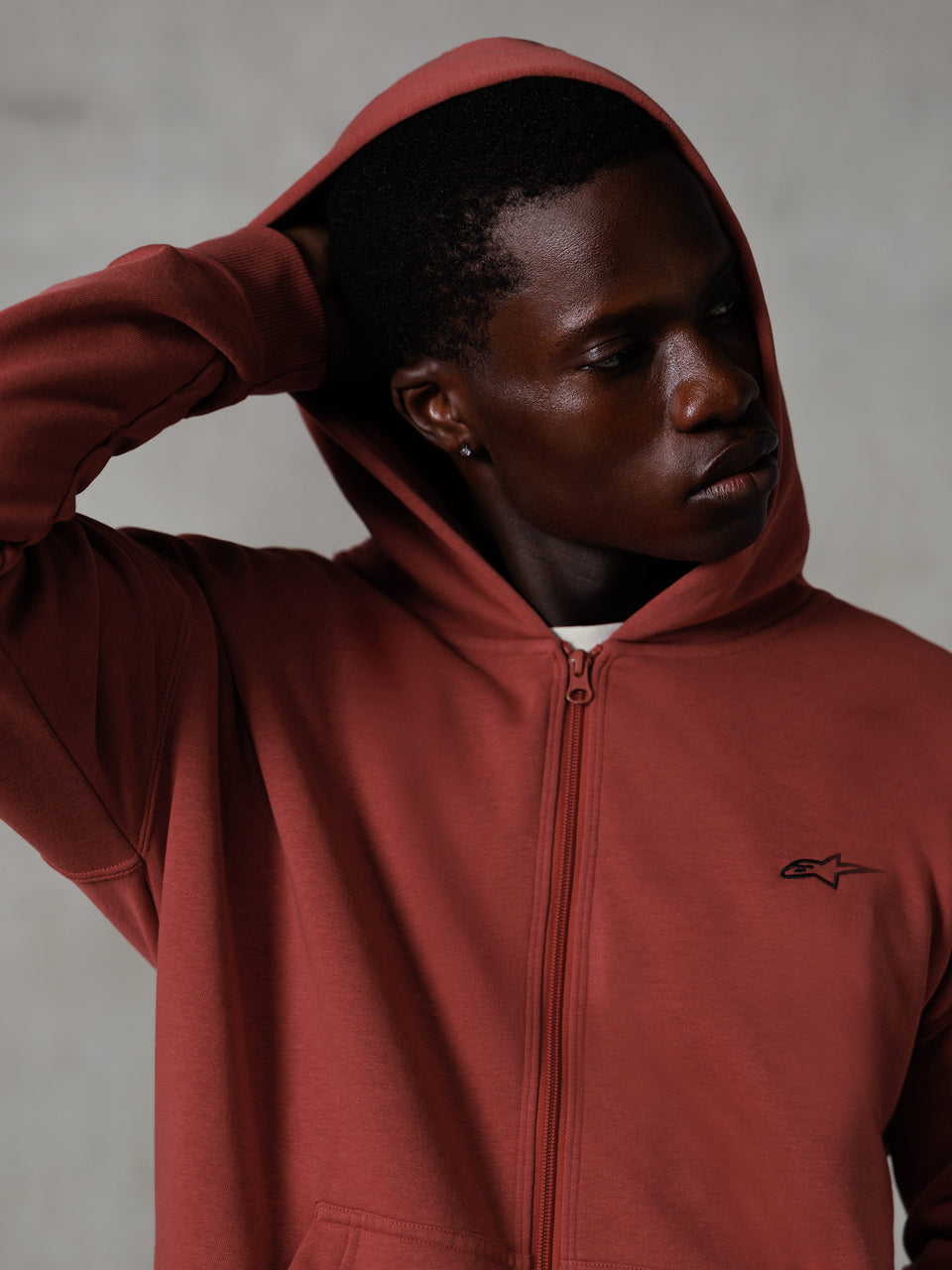 Alpinestars Essentials Ageless Zip Hoodie, vermelho tijolo, streetwear casual lifestyle, capuz levantado com a mão atrás da cabeça, pequeno logótipo Astars preto no peito, hoodie em fleece de corte descontraído