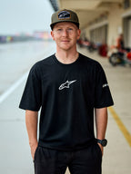 Homem a usar T-shirt de manga curta Alpinestars Frontal CSF, preta, logótipo Astars central branco, boné Alpinestars verde tropa, streetwear casual inspirado no desporto motorizado