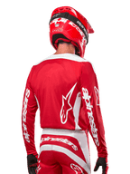 Piloto de motocross com camisola Alpinestars 2024 Fluid Lurv, em vermelho Mars e branco, vista traseira com logótipo Alpinestars branco nas costas e branding nas mangas, acompanhado por capacete vermelho e luvas pretas, equipamento profissional de corrida todo-o-terreno