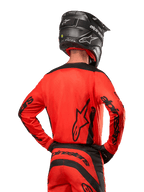 Piloto de motocross visto de costas a usar a camisola Alpinestars 2024 Fluid Lurv em Hot Orange e Preto, com marca Alpinestars em destaque nas costas e mangas, combinada com calças de motocross a condizer e um capacete preto, concebida para performance em competição off-road.