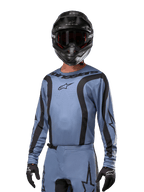Piloto de motocross a usar a camisola Alpinestars 2024 Fluid Lurv, azul claro e preto, combinada com capacete e óculos pretos, com detalhes gráficos pretos e o logótipo de estrela da Alpinestars, equipamento de performance para corridas todo-o-terreno