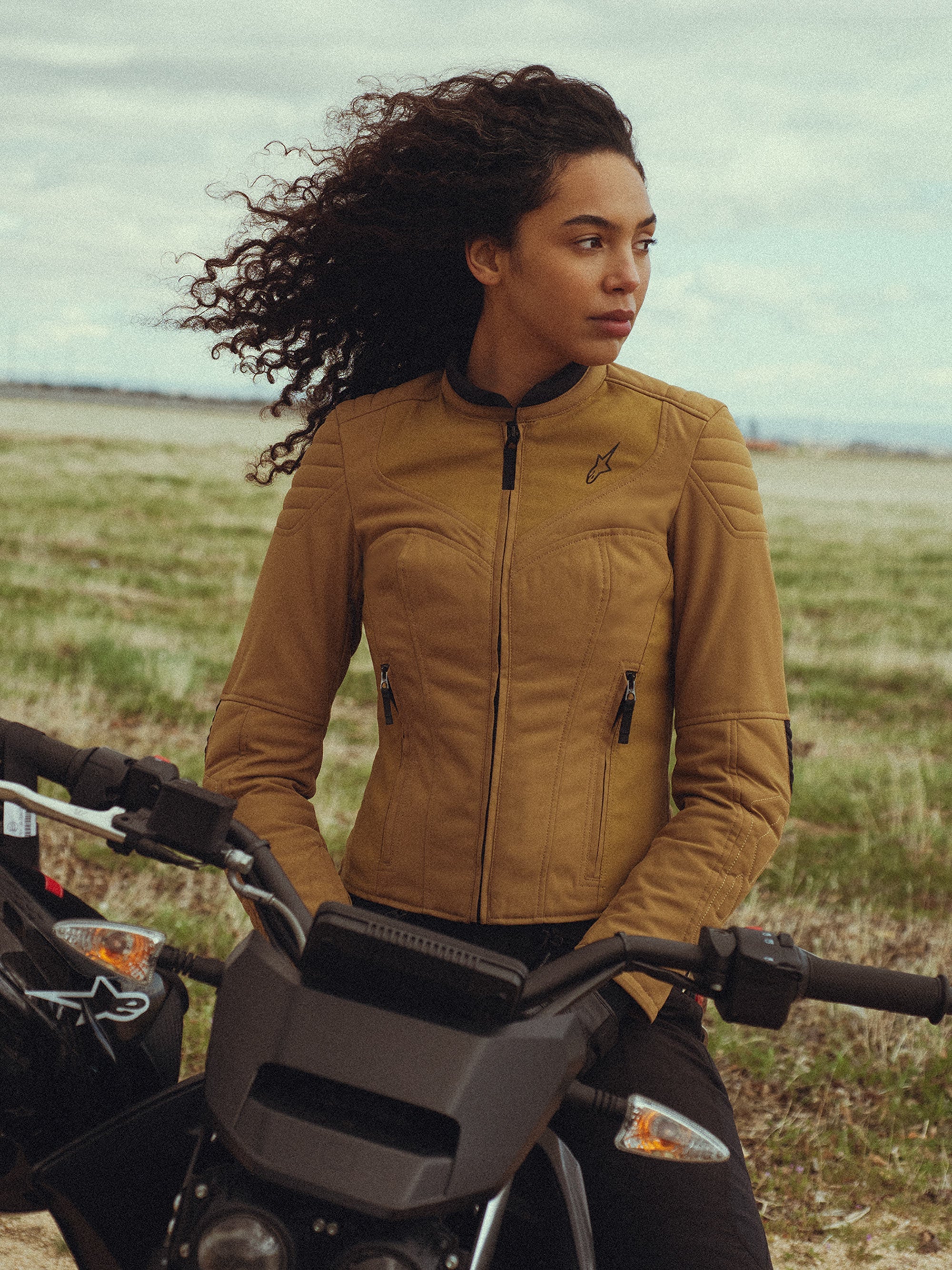 Alpinestars Isla WR Women's Jacket in Utility Brown Light Gold, worn by a woman sitting on a motorcycle in an outdoor field, featuring quilted shoulder details, a slim-fit silhouette, and urban contemporary styling.