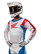 Motocross rider wearing the Honda Racer Iconic Jersey in White, Bright Blue, and Bright Red, featuring a large Honda wing logo on the chest, paired with a white Alpinestars helmet and matching white and black gloves, coordinated gear for off-road performance