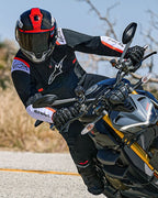 Motociclista inclinado numa curva numa moto naked, a usar o Casaco Alpinestars T-SPS Air v2 em preto com detalhes em branco e vermelho fluo, com capacete preto e vermelho e luvas pretas, equipamento têxtil de performance para condução urbana desportiva