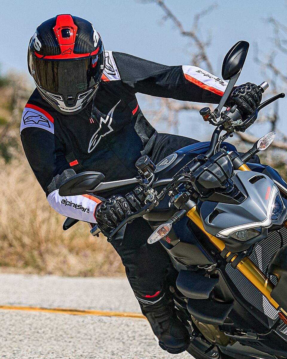 Motociclista inclinado numa curva numa moto naked, a usar o Casaco Alpinestars T-SPS Air v2 em preto com detalhes em branco e vermelho fluo, com capacete preto e vermelho e luvas pretas, equipamento têxtil de performance para condução urbana desportiva