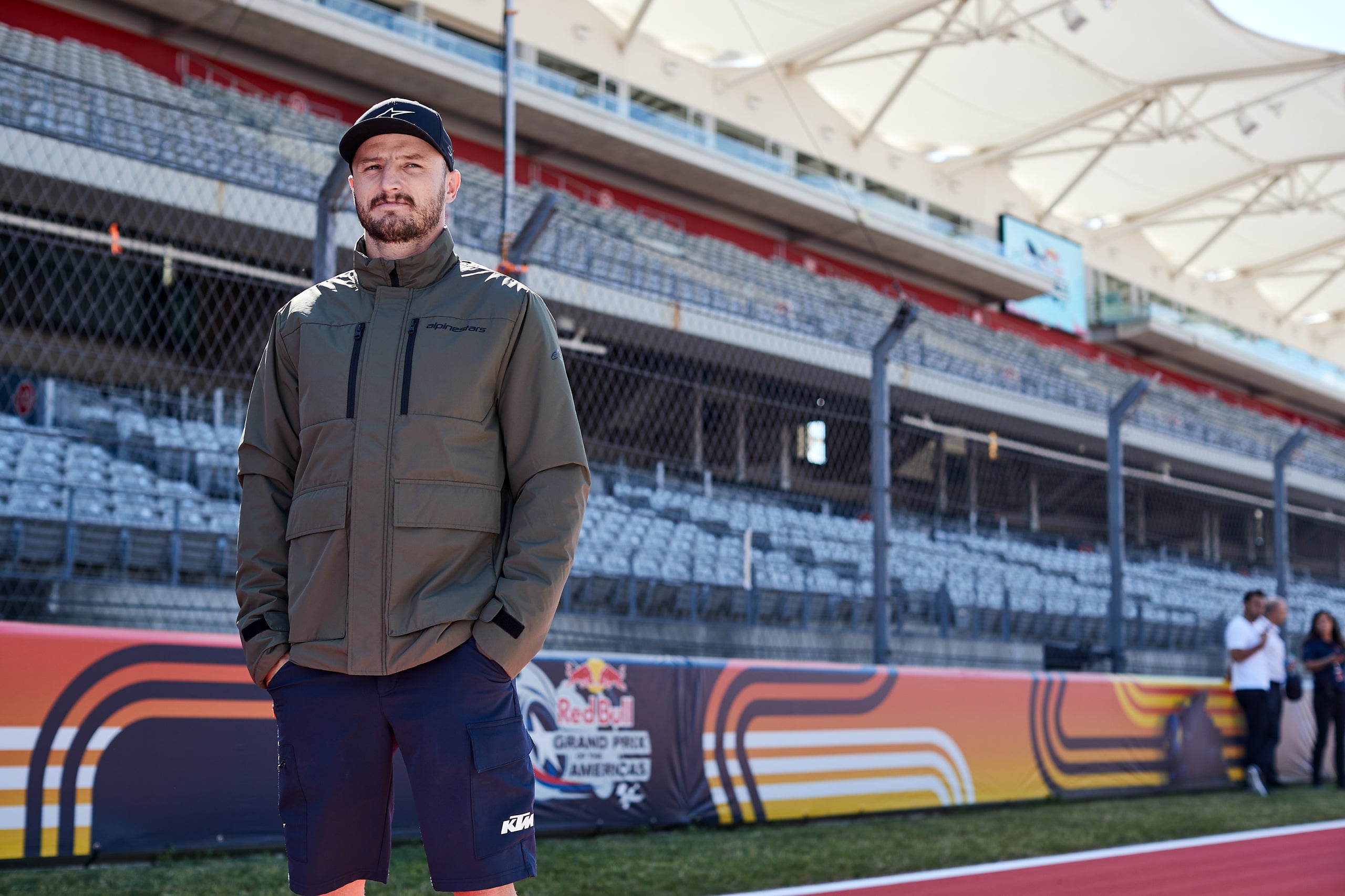 Jack Miller wearing a green Alpinestars jacket and cap standing on a race track with a blurred background
