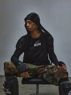 Alpinestars RSRV Nucleon SS Tee, t-shirt preta com logótipo branco RSRV, pessoa sentada com roupa urbana tática, calças camufladas verdes e castanhas, luvas sem dedos pretas, joelheiras pretas com proteção