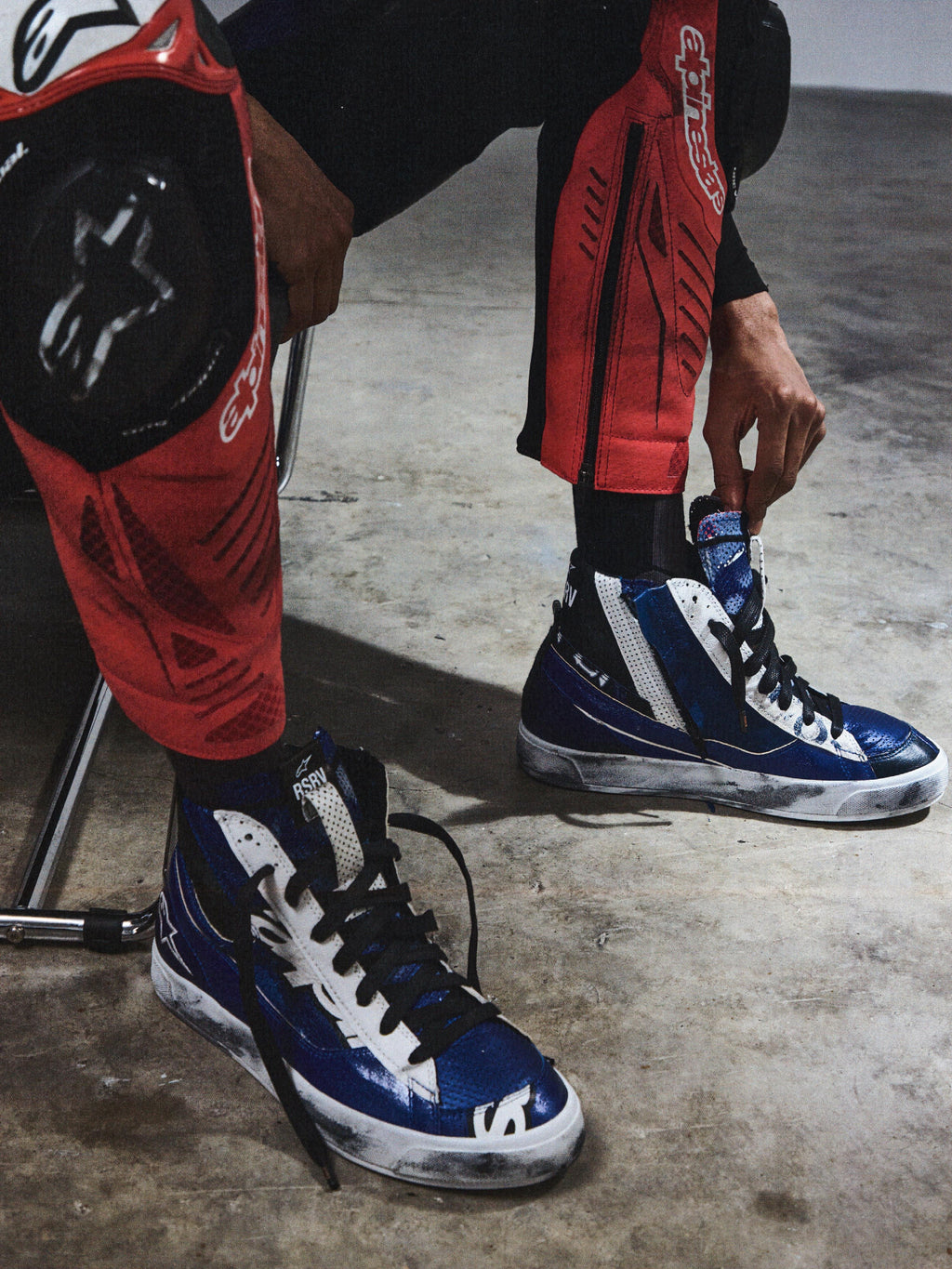 Alpinestars RSRV Legacy Shoe, sapatilhas altas azuis e brancas com atacadores pretos, estilo urbano casual, pessoa a ajustar as sapatilhas enquanto usa calças de proteção vermelhas e pretas, sola com desgaste visível, superfície de betão cinzento