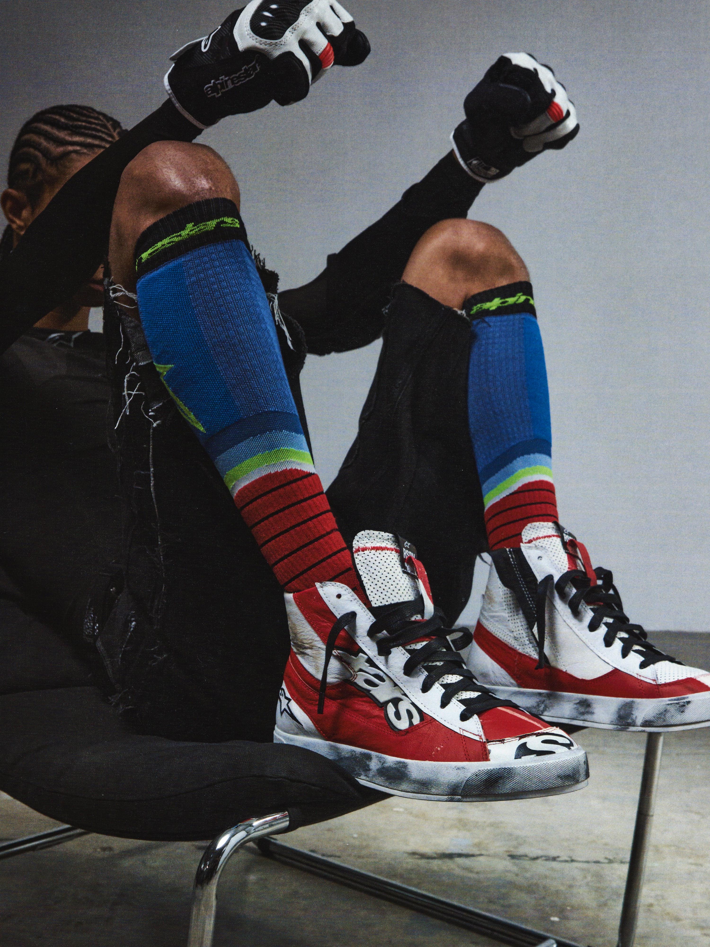 Alpinestars RSRV Legacy Shoe, sapatilhas altas vermelhas, brancas e pretas, estilo streetwear premium, pessoa sentada com meias coloridas às riscas, luvas pretas com detalhes vermelhos e brancos