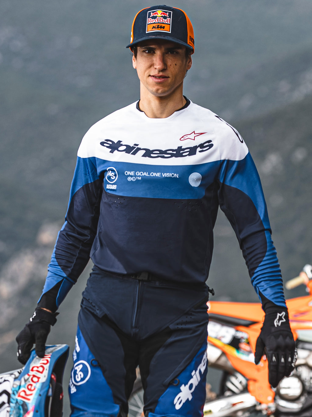 Piloto de motocross com camisola e calças Alpinestars Pro-Dura em azul marinho, azul brilhante e branco, com a marca 'One Goal One Vision', luvas pretas e a segurar um capacete de corrida, equipamento profissional de todo-o-terreno