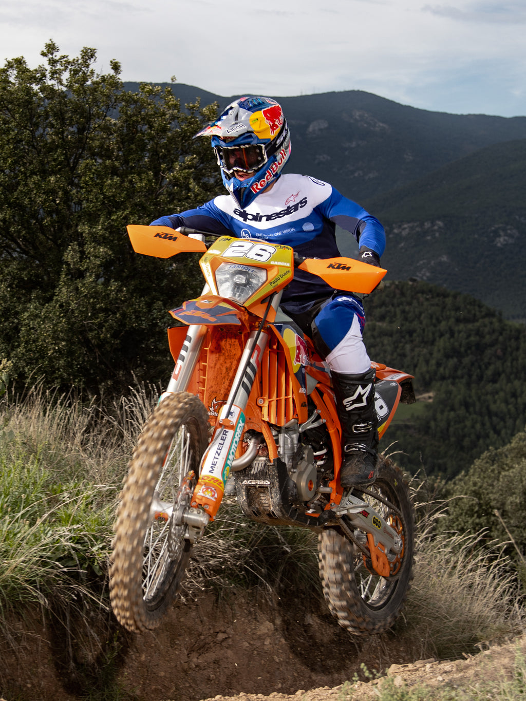 Piloto de motocross em ação numa moto KTM laranja, a usar o equipamento Alpinestars Pro-Dura em azul marinho, azul vivo e branco. Conjunto profissional de todo-o-terreno composto por camisola azul e branca e calças a condizer, usado com um capacete Red Bull e botas pretas num trilho de montanha.