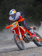 Motocross rider in action on an orange KTM bike, wearing Alpinestars Pro-Dura gear including a red, white, and black jersey and dark grey pants, red and white boots, and a Red Bull racing helmet, performing a sharp turn on a gravel trail