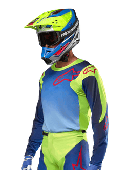 Alpinestars 2024 Racer Hoen Jersey, camisola de motocross, amarelo néon e azul marinho com logótipos vermelhos, usada com um capacete a condizer em amarelo, azul e vermelho e luvas azuis, equipamento vibrante para corridas off-road