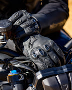 Luvas Alpinestars SP X 7, luvas de motociclismo em couro perfurado preto com protetores de articulações em TPU e deslizadores de dedos, mostradas nas mãos de um motociclista a segurar o guiador de uma mota.