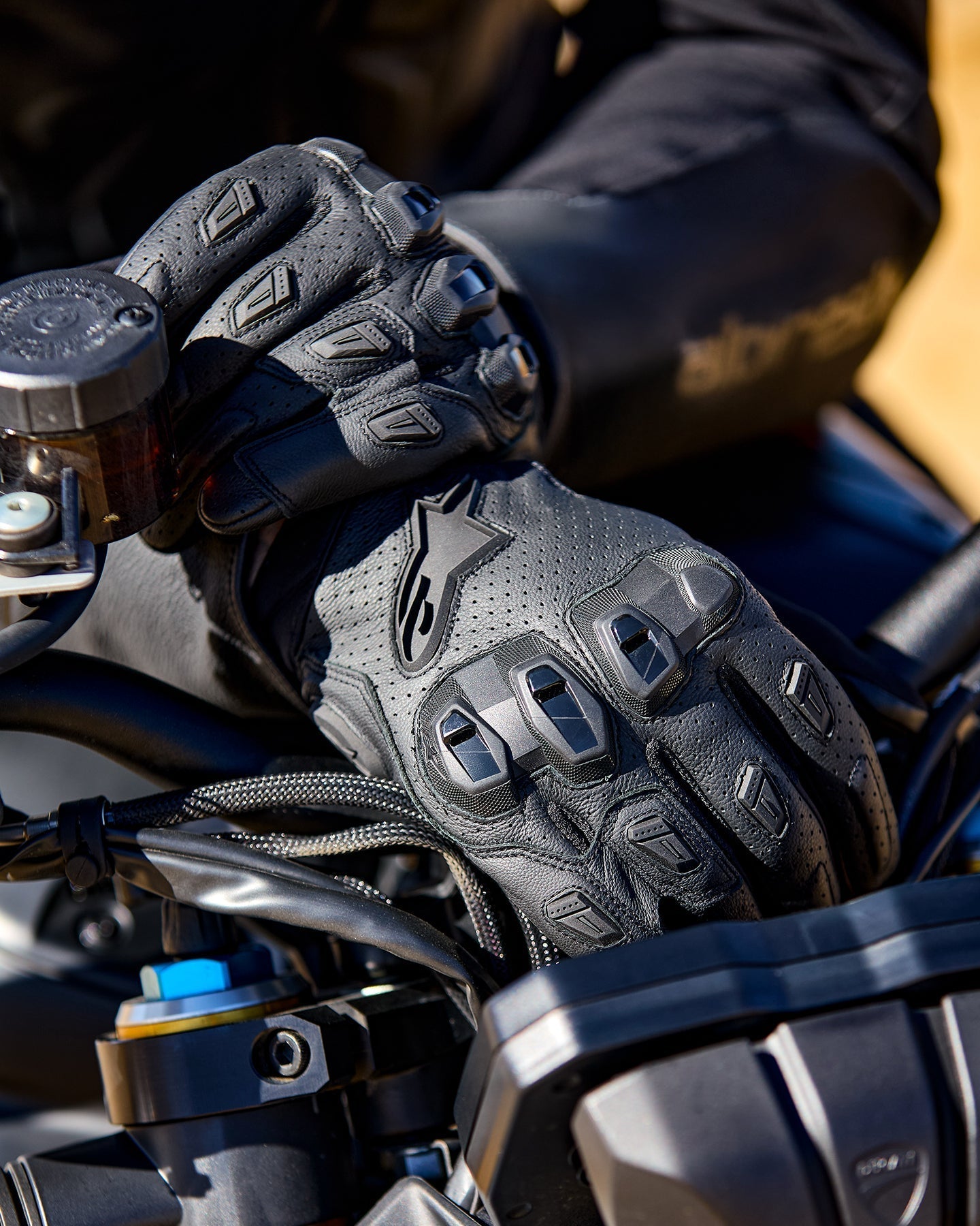 Luvas Alpinestars SP X 7, luvas de motociclismo em couro perfurado preto com protetores de articulações em TPU e deslizadores de dedos, mostradas nas mãos de um motociclista a segurar o guiador de uma mota.