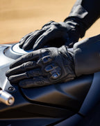 Luvas Alpinestars SP X 7, pretas, luvas de motociclismo de cano curto em couro perfurado, mãos apoiadas num depósito de mota escuro, com proteção visível nas articulações em TPU de dupla densidade e logótipo em relevo, concebidas para condução desportiva e de estrada.