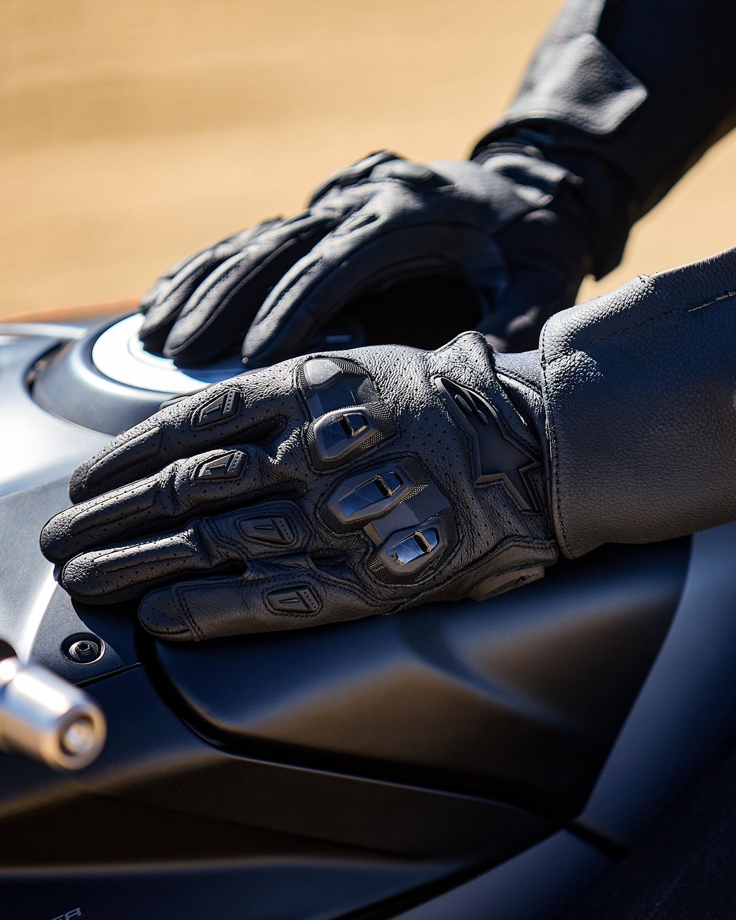Luvas Alpinestars SP X 7, pretas, luvas de motociclismo de cano curto em couro perfurado, mãos apoiadas num depósito de mota escuro, com proteção visível nas articulações em TPU de dupla densidade e logótipo em relevo, concebidas para condução desportiva e de estrada.