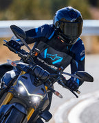 Motorcycle rider leaning into a curve wearing the Alpinestars T-Stunt Air Jacket in Black, Dark Blue, and Bright Blue, paired with a black helmet and gloves, action shot on a paved road, performance sport riding gear