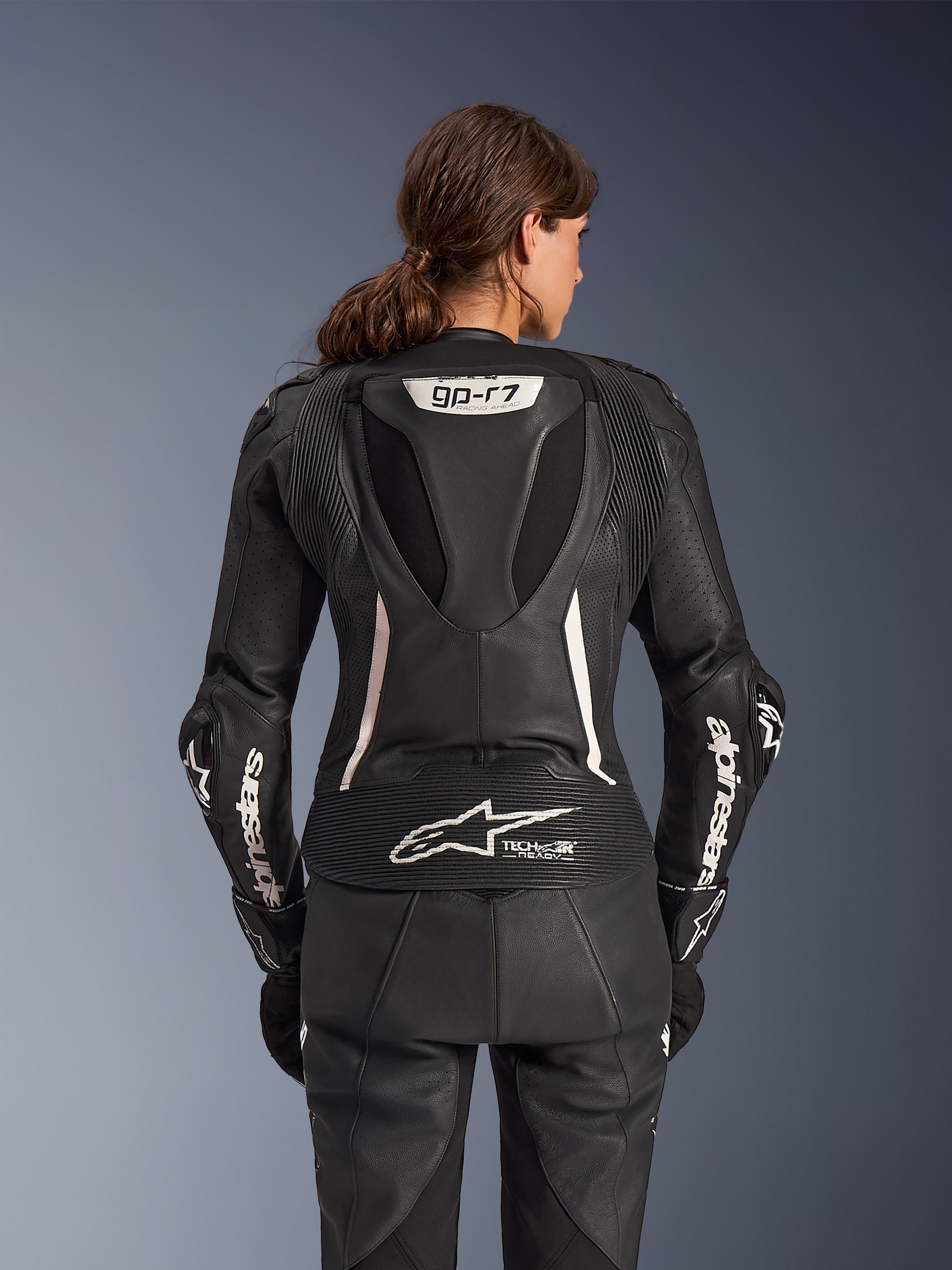 Fato de Cabedal de 2 Peças Alpinestars Stella GP-R7, fato de motociclismo para mulher, preto com detalhes brancos, vista traseira mostrando a corcova aerodinâmica Supertech R10 com o logótipo GP-R7, marcação Tech-Air Ready na parte inferior das costas, lettering Alpinestars branco nas mangas e painéis elásticos em acordeão para performance de corrida