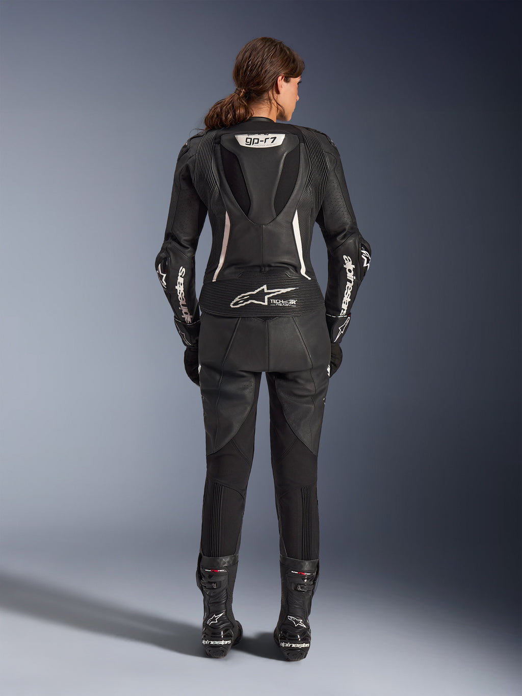 Fato de Cabedal de 2 Peças Alpinestars Stella GP-R7, fato de motociclismo para mulher, preto com detalhes brancos, vista traseira com a corcova aerodinâmica GP-R7 e a marca Tech-Air Ready, logótipos Alpinestars brancos nas mangas, painéis extensíveis em acordeão, usado com botas de corrida pretas