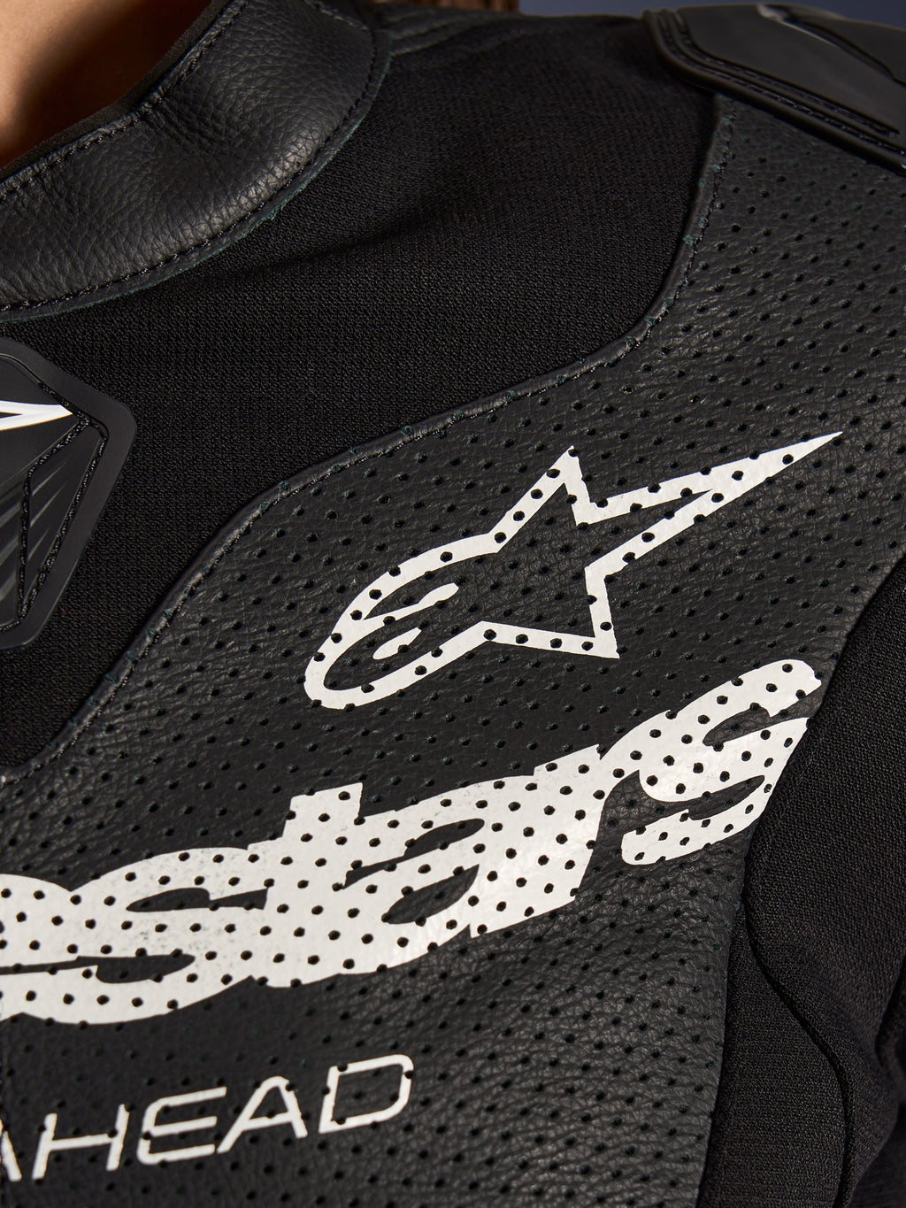 Pormenor do Fato de Cabedal Stella GP-R7 de 2 Peças da Alpinestars em preto, com painéis de pele perfurada, um logótipo Alpinestars branco no peito e secções de tecido elástico respirável.
