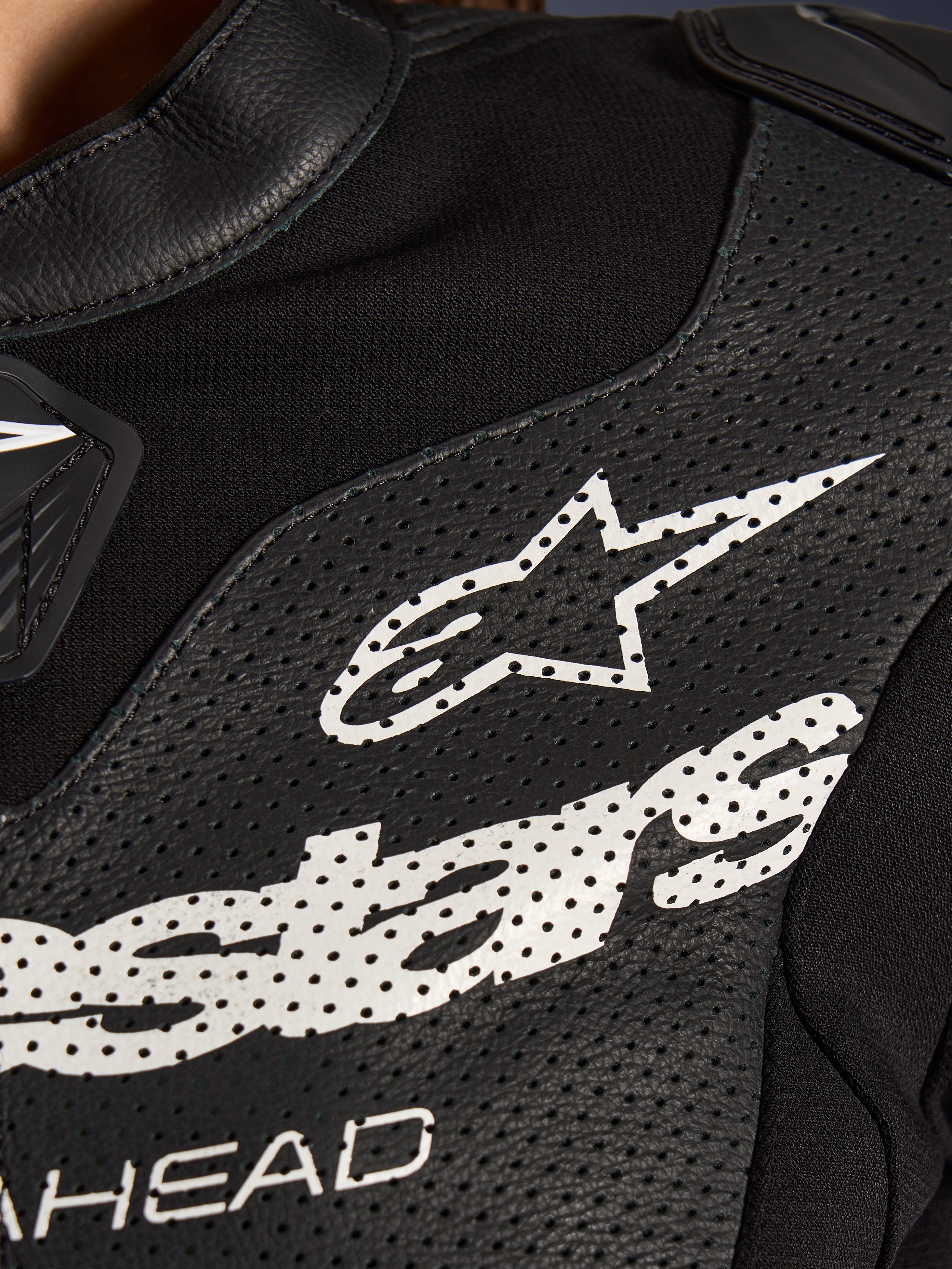 Pormenor do Fato de Cabedal Stella GP-R7 de 2 Peças da Alpinestars em preto, com painéis de pele perfurada, um logótipo Alpinestars branco no peito e secções de tecido elástico respirável.