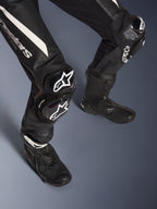 Detalhe das pernas do Fato de Cabedal Stella GP-R7 de 2 Peças da Alpinestars, preto com detalhes brancos, com proteção de joelho e deslizadores GP DFS RR com a marca Alpinestars, combinado com botas de corrida pretas, concebido para performance em pista