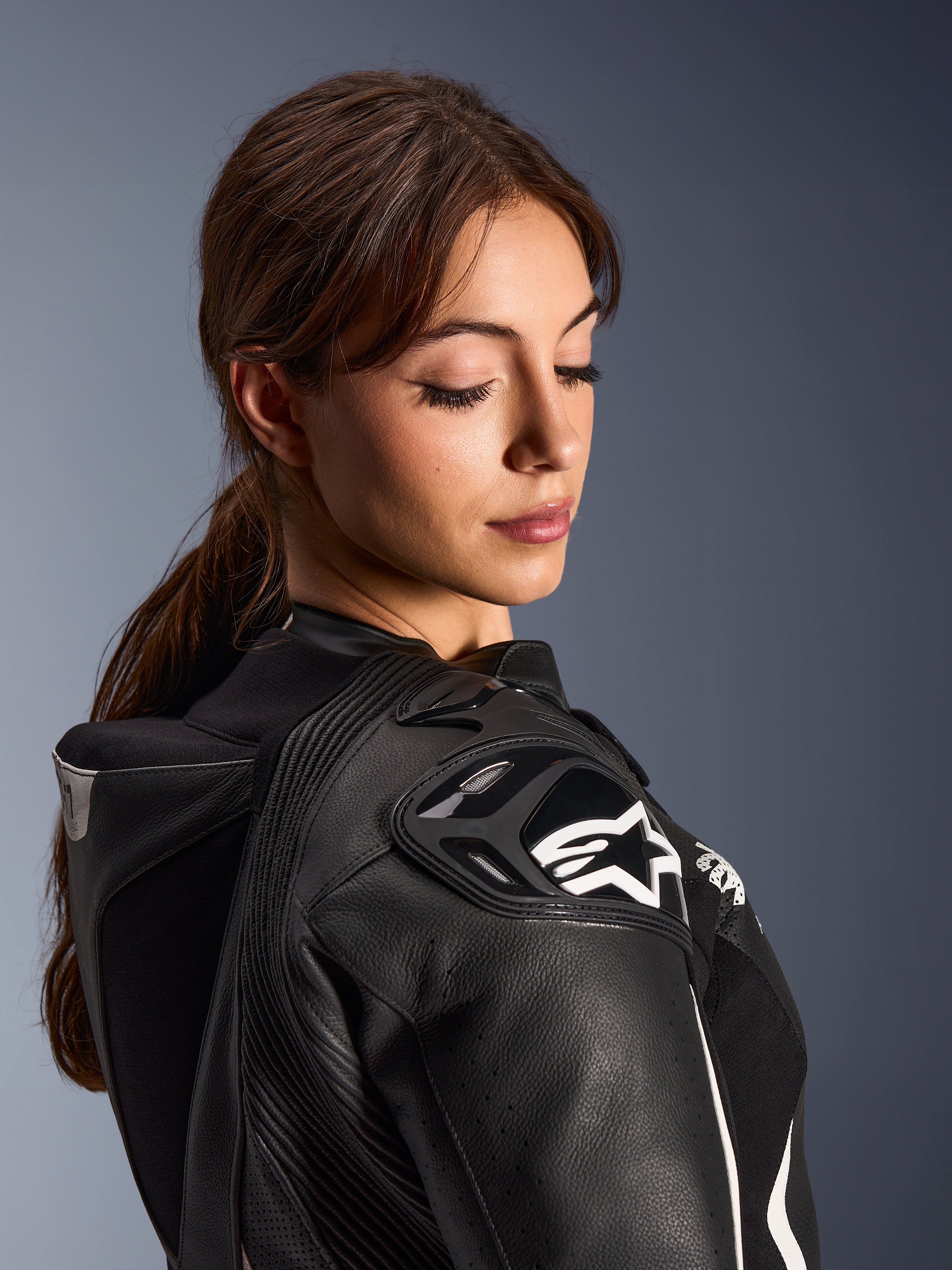 Mulher a usar o Fato de Cabedal de 2 Peças Alpinestars Stella GP-R7, vista em grande plano do ombro em pele preta com proteção DFS RR e logótipo de estrela branca, apresentando a corcova aerodinâmica integrada e detalhes perfurados.