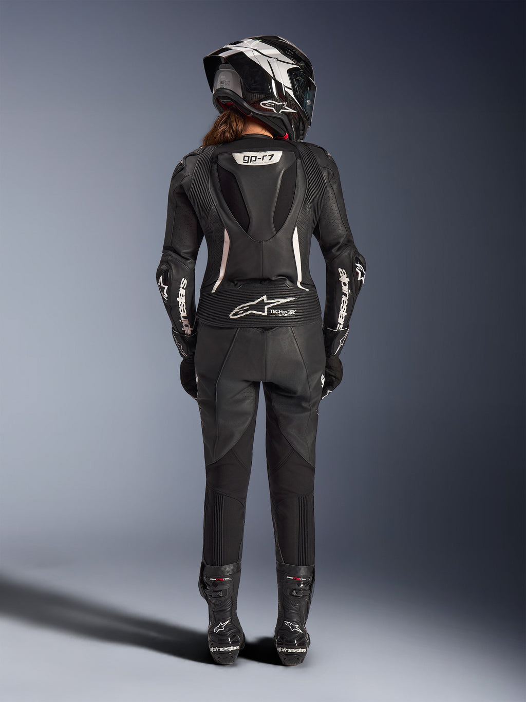 Fato de Cabedal de 2 Peças Alpinestars Stella GP-R7 Feminino, preto com detalhes brancos, vista traseira que mostra a corcova aerodinâmica Supertech R10 e o logótipo Tech-Air Ready, usado com capacete integral, luvas de corrida e botas de cabedal a condizer para condução em pista e desportiva.