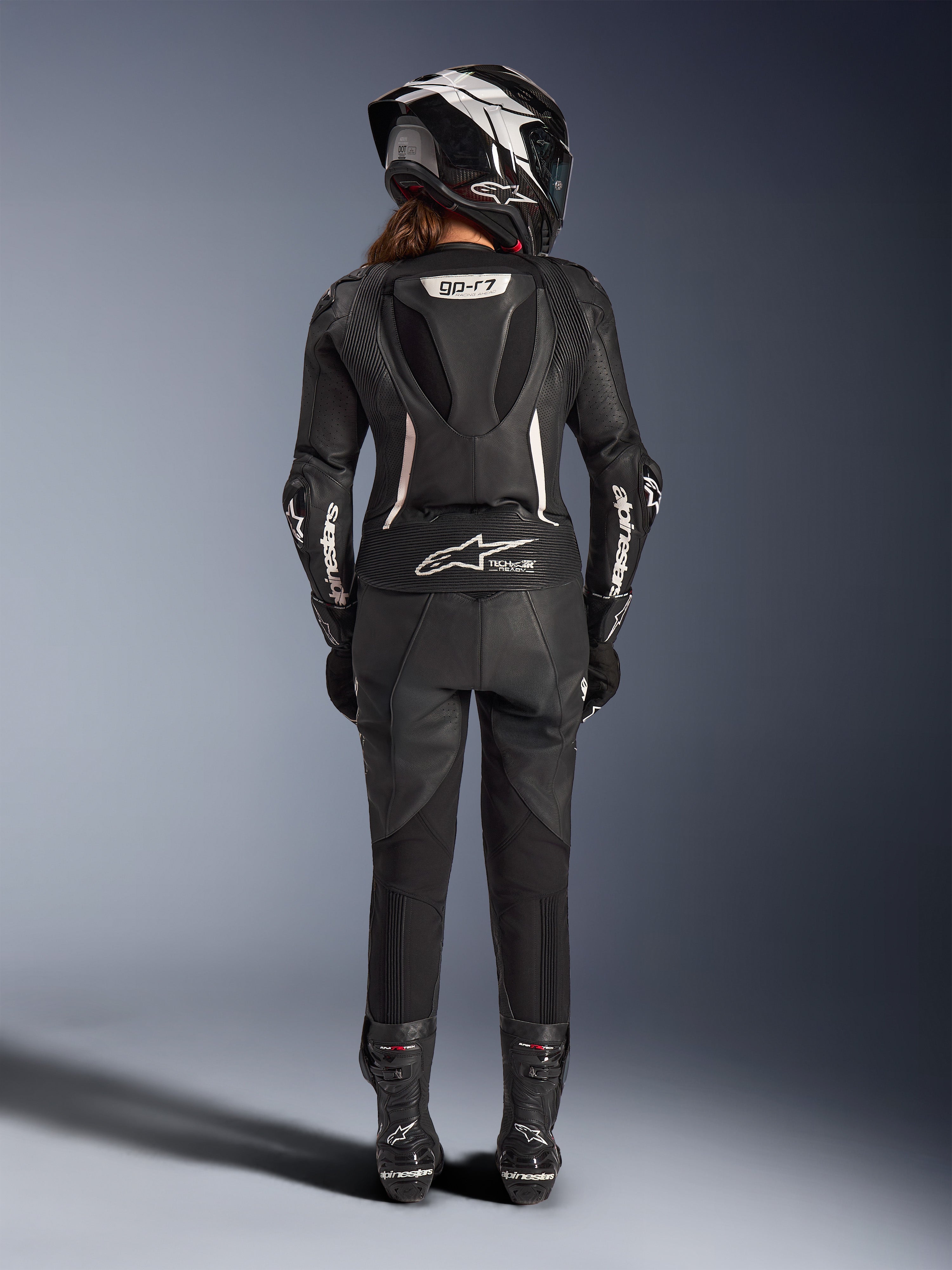 Fato de Cabedal de 2 Peças Alpinestars Stella GP-R7 Feminino, preto com detalhes brancos, vista traseira que mostra a corcova aerodinâmica Supertech R10 e o logótipo Tech-Air Ready, usado com capacete integral, luvas de corrida e botas de cabedal a condizer para condução em pista e desportiva.