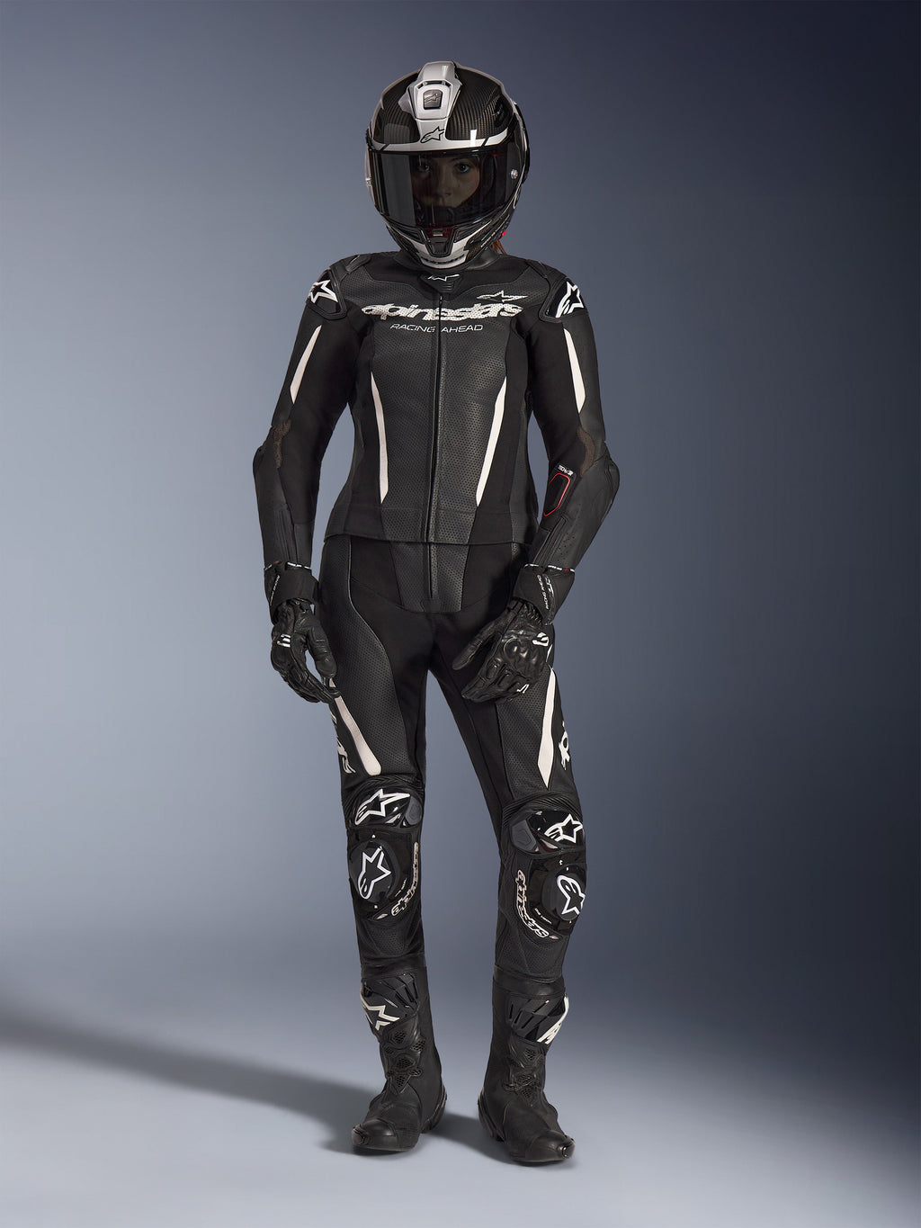 Motociclista feminina com o fato de cabedal de 2 peças Alpinestars Stella GP-R7 em preto com detalhes brancos, capacete integral de fibra de carbono, luvas de corrida e botas pretas, apresentando painéis de pele perfurada e armadura de proteção integrada, equipamento profissional para performance em pista e estrada