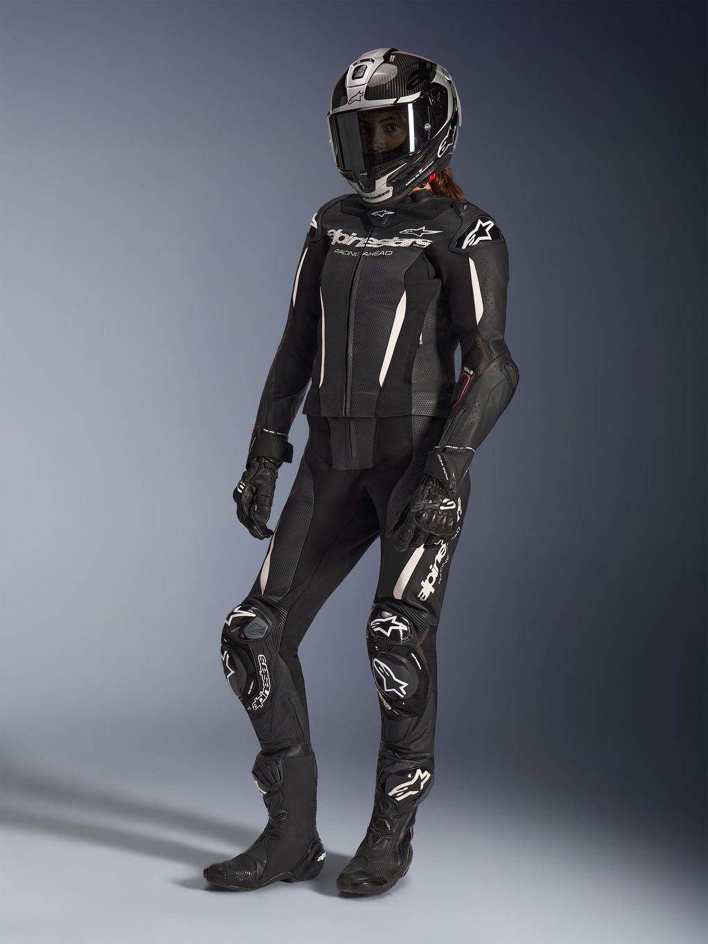 Alpinestars Stella GP-R7 2-Piece Leather Suit, fato de motociclismo para mulher, preto com detalhes brancos, usado por uma motociclista com capacete integral de corrida, luvas pretas e botas, com painéis de pele perfurada e logótipo Alpinestars branco, equipamento de corrida profissional e de performance desportiva