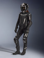 Alpinestars Stella GP-R7 2-Piece Leather Suit, fato de motociclismo para mulher, preto com detalhes brancos, usado por uma motociclista com capacete integral de corrida, luvas pretas e botas, com painéis de pele perfurada e logótipo Alpinestars branco, equipamento de corrida profissional e de performance desportiva