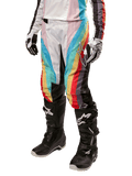 Calças Alpinestars Stella Techstar 2024 para Mulher, calças de motocross, pretas multicor com riscas diagonais vermelhas, amarelas e azuis, usadas com botas de condução pretas e luvas brancas, com painéis de proteção pretos nos joelhos e logótipo de destaque para corridas de todo-o-terreno.