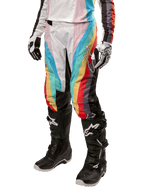 Calças Alpinestars Stella Techstar 2024 para Mulher, calças de motocross, pretas multicor com riscas diagonais vermelhas, amarelas e azuis, usadas com botas de condução pretas e luvas brancas, com painéis de proteção pretos nos joelhos e logótipo de destaque para corridas de todo-o-terreno.