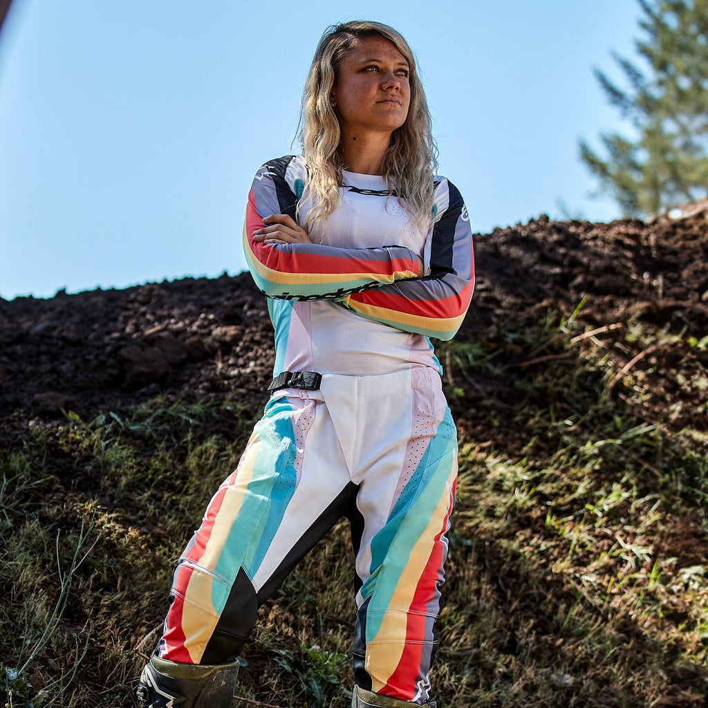 Piloto de motocross feminina com calças Alpinestars Stella Techstar Femininas 2024 e camisola a condizer, design em Preto Multicolor com base branca e riscas vibrantes em azul, amarelo e vermelho, de pé com os braços cruzados numa pista de terra, equipamento profissional de todo-o-terreno