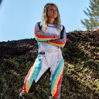 Piloto de motocross feminina com calças Alpinestars Stella Techstar Femininas 2024 e camisola a condizer, design em Preto Multicolor com base branca e riscas vibrantes em azul, amarelo e vermelho, de pé com os braços cruzados numa pista de terra, equipamento profissional de todo-o-terreno