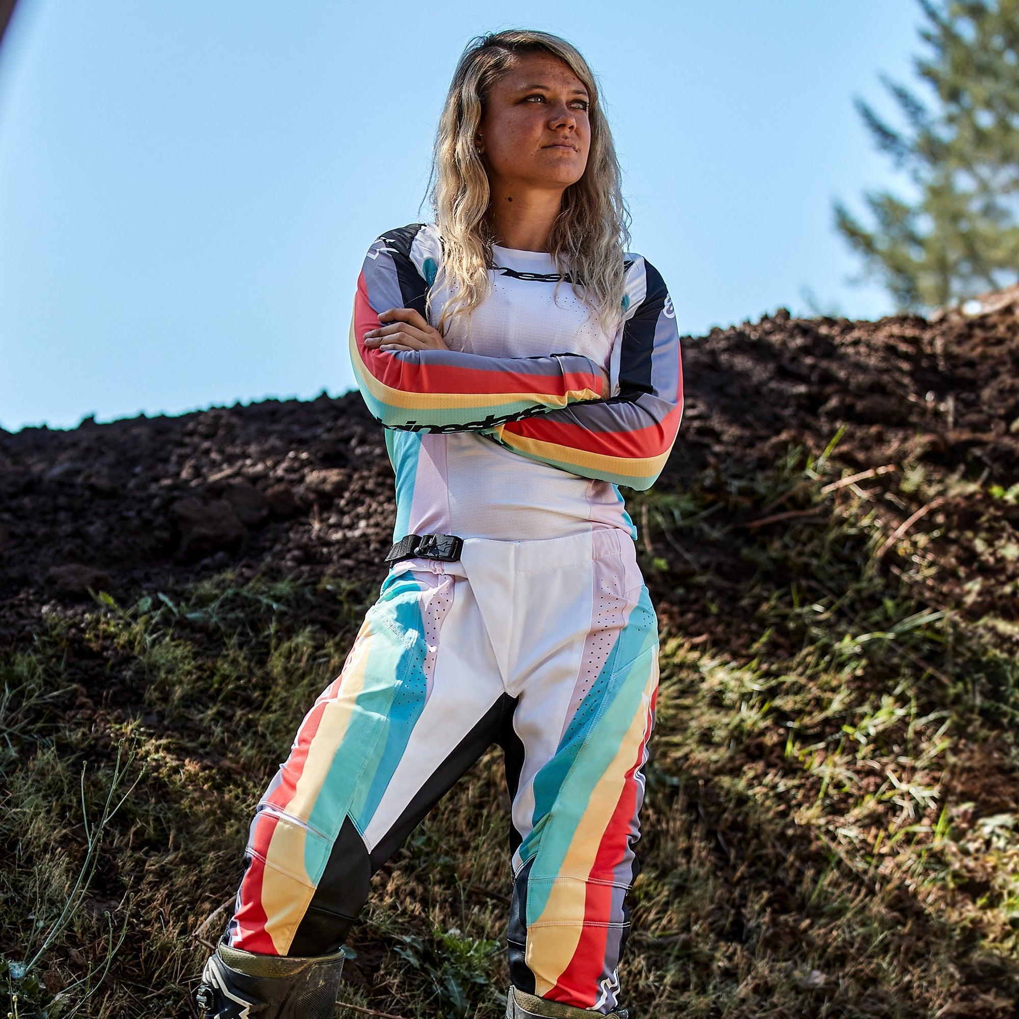 Piloto de motocross feminina com calças Alpinestars Stella Techstar Femininas 2024 e camisola a condizer, design em Preto Multicolor com base branca e riscas vibrantes em azul, amarelo e vermelho, de pé com os braços cruzados numa pista de terra, equipamento profissional de todo-o-terreno