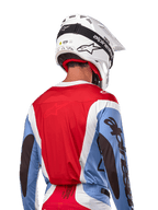 Camisola Alpinestars Techstar Ocuri 2024, camisola de motocross, azul claro, vermelho Mars e branco, vista traseira do piloto com capacete branco e óculos pretos, apresenta logótipos pretos de destaque nas mangas e corte ergonómico de corrida para performance em todo-o-terreno.