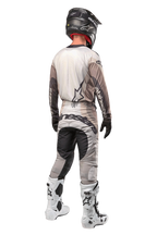 Alpinestars 2024 Techstar Pneuma Pants, calças de motocross, Areia Escura e Cinzento Iron Dust, vista traseira usada por um piloto com uma camisola a condizer, botas brancas e pretas e um capacete preto, com logótipo Alpinestars proeminente no assento e painéis escuros reforçados