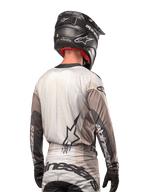 Piloto de motocross visto de trás a usar a camisola Alpinestars Techstar Pneuma 2024 em Dark Sand Iron Dust Gray, combinada com um capacete preto com acabamento em carbono, com um logótipo de estrela grande nas costas e a marca Alpinestars nas mangas.