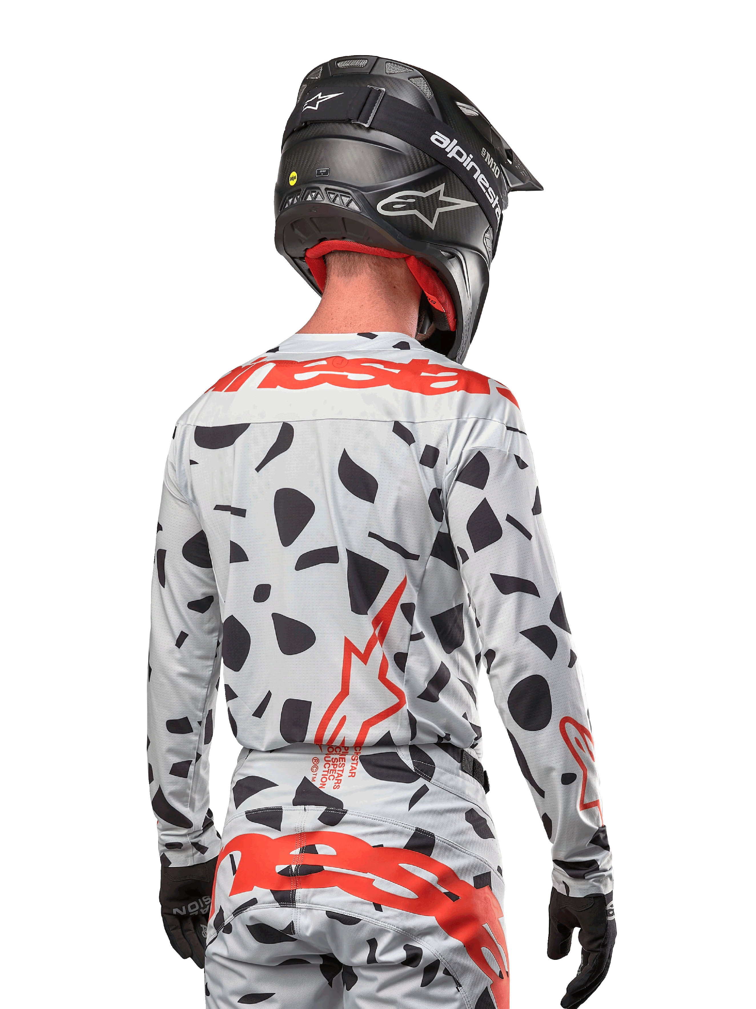 Piloto de motocross visto de costas a usar a camisola Alpinestars 2024 Techstar Rantera em Haze Gray Camo, com uma base cinza clara e manchas gráficas pretas, logótipos Alpinestars em vermelho vibrante nas costas e ombros, acompanhado por calças a condizer e capacete preto.