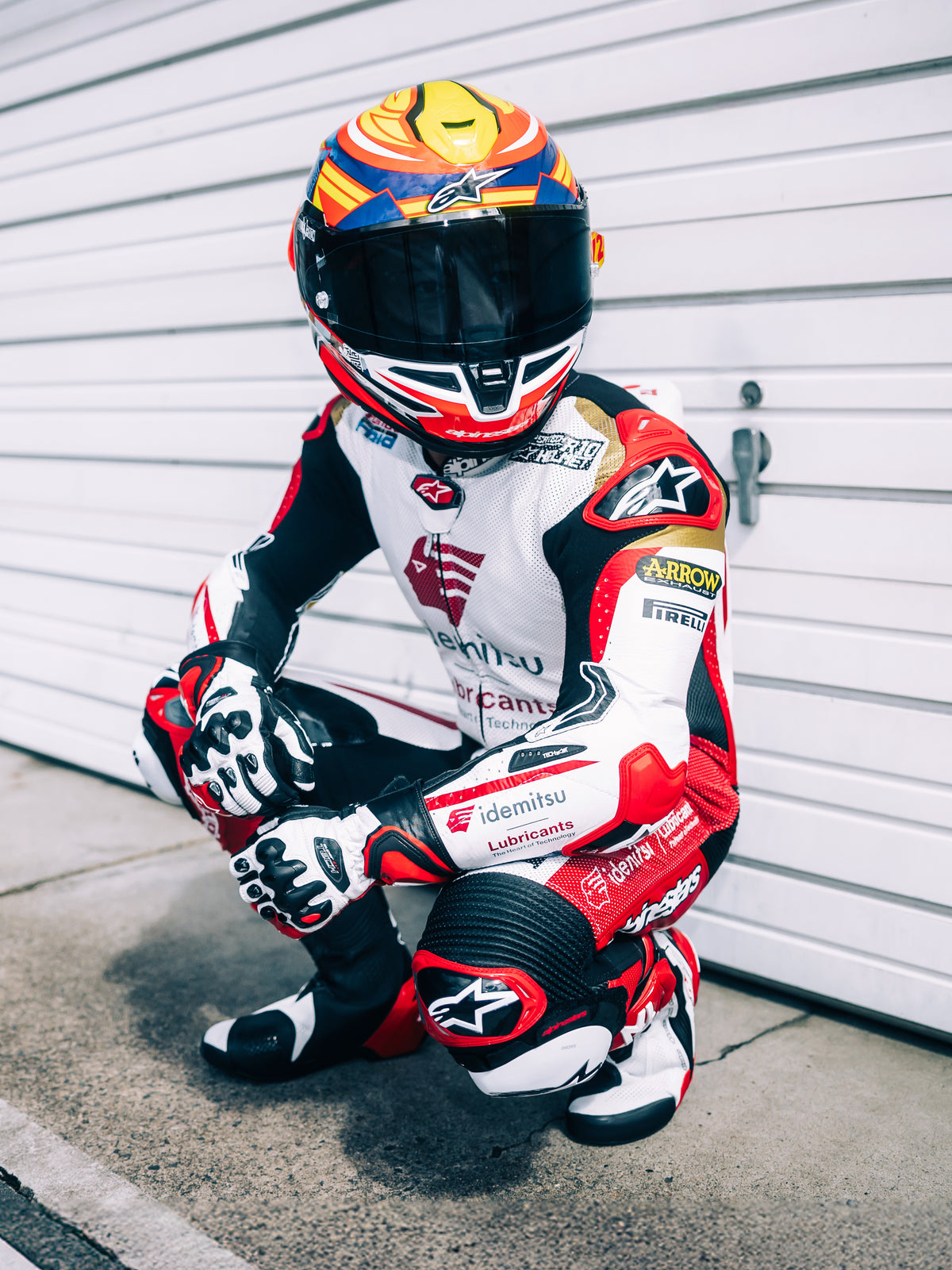 Capacete Alpinestars S-R7 Taiyo Furusato, capacete integral de moto para corrida, vermelho, branco, azul e amarelo fluorescente, certificado ECE 22.06, usado por piloto de moto com macacão de couro e viseira fumada.