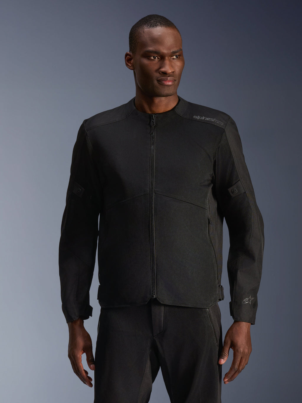 Modelo masculino a usar Casaco Alpinestars C-1 Air em Preto, casaco de motociclismo contemporâneo com amplos painéis de malha técnica e fecho de correr frontal integral, combinado com calças de condução pretas a condizer, estética urbana e de deslocações diárias